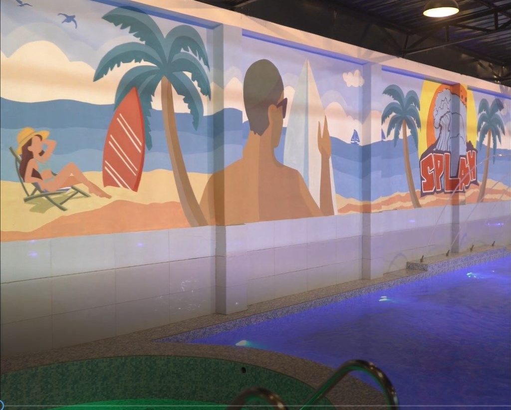 An Nexora Structure Swimming Pool Area With A Large Beach-Themed Wall Mural Featuring Sunbathers, Surfboards, And Palm Trees Under Bright Overhead Lighting.